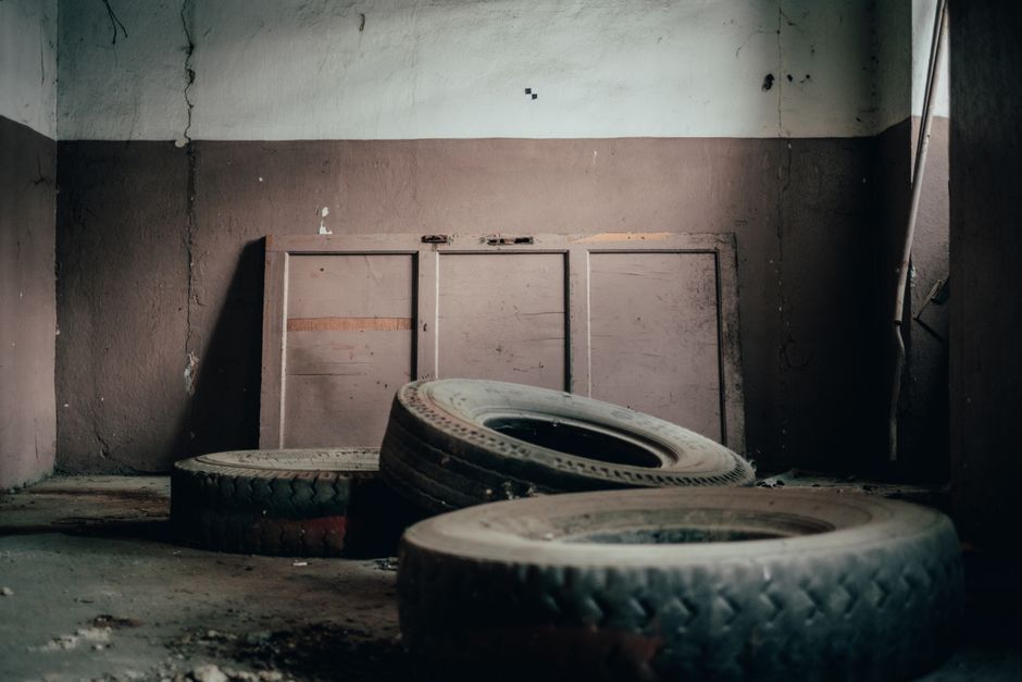 Inside an abandoned, dimly lit room with peeling paint and cracked walls, three used vehicle tires with worn rubber and visible tread patterns rest on a dusty concrete floor. The tires, varying in size, are positioned in a cluster towards the foreground, with the largest tire leaning slightly against another. Behind them, a set of old, boarded-up wooden panels or doors, painted in a faded beige or light brown, are mounted against the wall. The wall has patches of chipped paint and minor surface damage, contributing to the overall neglected appearance. To the right, a metal rod or pole is partially visible, leaning or hanging against the wall, and the environment lacks natural light, emphasizing a somber, abandoned atmosphere. This scene could be associated with private waste collection or on-site clearance activities, often undertaken by waste removal services such as Waste Clearance Lewisham to manage discarded tires and automotive waste in similar settings.