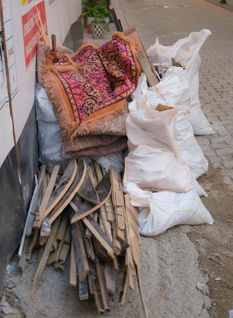 A pile of discarded household items and rubbish placed on a sidewalk outside a building. The collection includes a rolled-up, patterned area rug with shades of pink, purple, and orange, resting on top of what appears to be a larger, muted-colored carpet or fabric. Next to the rugs, there are several large white sacks filled with unknown materials, some tied at the top and slightly bulging. Leaning against the sacks and partially stacked on the ground are pieces of wooden furniture or construction debris, including bent and broken wooden slats or trim with a light, natural finish. The scene is set against a wall with a red and white sign, possibly indicating a nearby property or safety instructions. The surrounding environment includes a paved walkway with visible paving stones and a section of street or driveway visible in the background, suggesting an outdoor rubbish removal scene. The scene appears to be part of an independent waste collection or on-site clearance arranged by Waste Clearance Lewisham as part of a rubbish removal service, illustrating a typical example of bulk rubbish awaiting collection outside a property.