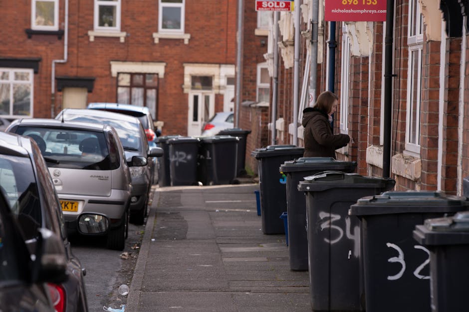 A daytime street scene in a residential area featuring a row of dark grey wheelie bins lined along the pavement outside brick houses with white window frames and stone sill details. The bins are positioned close to the buildings, with some lids closed and others slightly ajar, and are marked with white spray-painted numbers. A woman dressed in a dark brown coat is standing next to one of the bins, appearing to be in the process of disposing of waste. Several parked cars, including a silver hatchback and a dark vehicle with visible rear window and side mirror, are situated along the curb on the left side of the image. The background shows more parked cars and additional houses with brick facades, some displaying 'sold' signs in the windows. The setting appears to be an urban street scene that highlights private waste disposal options, with a focus on rubbish collection outside residential properties, relevant to waste clearance and rubbish removal services.