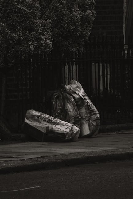 At the edge of a paved street, three large black plastic rubbish bags filled with waste are stacked against a dark wooden fence, with a metal gate visible behind them. The bags appear tightly packed and are slightly reflective, with some creases and folds indicating crumpled material inside. To the left of the rubbish bags, a small section of a tree or bush with dense, dark foliage is visible, adding a natural element to the scene. The environment suggests an urban or residential area, possibly near a driveway or alleyway, where private waste collection might take place. The overall lighting is subdued, creating a shadowed atmosphere, consistent with late evening or overcast weather. This scene relates to independent or private rubbish clearance services, such as those offered by Waste Clearance Lewisham, which may handle on-site waste removal and disposal outside standard council collection systems.