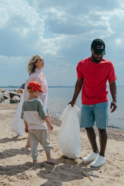 A man in a red T-shirt, denim shorts, and a black cap with a logo is standing on a sandy beach, holding a white rubbish bag in his right hand and assisting in collecting waste. Behind him, a woman with curly blonde hair, wearing a light pink dress, stands near the water's edge, looking toward the man and the children. Two young children, one wearing a red cap with a pattern, a beige and green shirt, and beige shorts, are also present, each holding white rubbish bags, participating in a beach clean-up. The background features a calm body of water, a rocky shoreline, and a partly cloudy sky, indicating a natural outdoor environment. The scene is well-lit with natural daylight, emphasizing a peaceful, active effort in waste collection in an outdoor setting often associated with leisure and recreation, aligning subtly with private or independent rubbish removal activities facilitated by waste clearance services. Waste Clearance Lewisham specializes in rubbish removal and environmental cleanup tasks, supporting local efforts to maintain clean spaces without relying solely on municipal collection services.