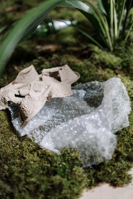 Close-up view of broken polystyrene packaging and a piece of flattened paperboard carton discarded on a bed of green moss outdoors. The polystyrene exhibits a textured, bubbly surface with irregular edges and transparent appearance, contrasting with the matte, slightly rough texture of the paperboard, which is light beige in color. Behind the debris, there are large, broad green leaves with smooth surfaces and pointed tips, suggesting a garden or natural setting. The moss surrounding the rubbish is dense, soft, and vibrant, adding a natural backdrop that emphasizes the man-made waste. The scene is illuminated by natural daylight, creating subtle shadows and highlights on the textured surfaces of the packaging and moss. This visual captures components typical of waste encountered during domestic or garden clearance, aligning with the services of waste clearance specialists like Waste Clearance Lewisham, who handle various rubbish types through private collection and on-site clearance methods.