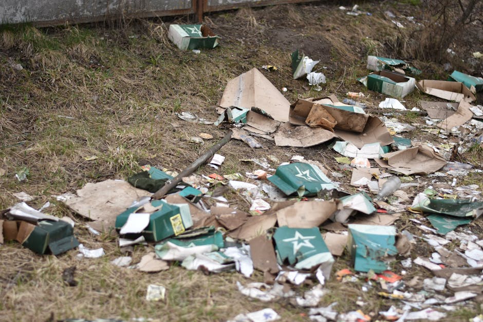 An outdoor scene showing scattered discarded cardboard boxes, many flattened or torn, along with crumpled paper and plastic packaging strewn across a patch of uneven grass and soil. Some boxes are partially collapsed, revealing torn edges and exposed corrugated layers in shades of brown, green, and white. A few boxes display printed logos or branding details, indicating they once contained goods or products. The debris extends from a grassy area near a concrete or brick structure at the top of the image, with small pieces of miscellaneous rubbish such as paper, plastic wrappers, and fragments of packaging visible among the larger waste items. The environment appears untidy, typical of waste left behind for cleanup or disposal, representing typical conditions for private rubbish collection or on-site clearance services offered by waste management companies such as Waste Clearance Lewisham. The natural light highlights the textures of the rubbish and the uneven terrain, emphasizing the importance of proper waste removal and environmental care.