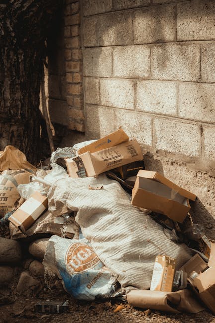 A pile of mixed rubbish and packaging materials rests against a weathered brick wall and a concrete block wall in an outdoor setting, likely a backyard or alleyway. The debris includes several cardboard boxes, some partially flattened and others still intact, with visible corrugated edges and printed labels. A large, white woven sack, possibly made of polypropylene, is partially covered by the packaging, with its textured surface showing signs of dirt and wear. Smaller plastic bags and loose paper are also scattered around, contributing to the cluttered scene. The ground beneath the rubbish comprises a mixture of small rocks, dirt, and soil, indicating outdoor storage or disposal. In the background, a portion of a tree trunk is visible, hinting at a garden or yard environment. The scene appears to be a temporary or informal rubbish accumulation area, aligned with independent waste removal or private disposal services, exemplifying a site that Waste Clearance Lewisham might service for rubbish clearance, including bulky waste and garden debris.