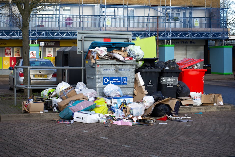A large collection of overflowing rubbish and waste, including various black and black-and-white plastic trash bags, flattened cardboard boxes, and miscellaneous packaging, piled near a metal railing on a paved urban sidewalk. Prominent in the scene is a gray mixed paper and cardboard recycling bin with its lid open, spilling loose paper and packaging onto the ground. Surrounding the main rubbish heap are additional black rubbish bins, one of which is red, and some boxed items lying on the pavement. In the background, there is a building under construction covered with blue safety scaffolding, along with shopfronts and signs for local businesses. A parked car is partially visible on the left side, with trees lining the street. The scene appears to be in a busy commercial area, illustrating unsalvaged waste that might relate to private rubbish disposal or unauthorized dumping, which waste clearance services like Waste Clearance Lewisham could help to manage more sustainably.
