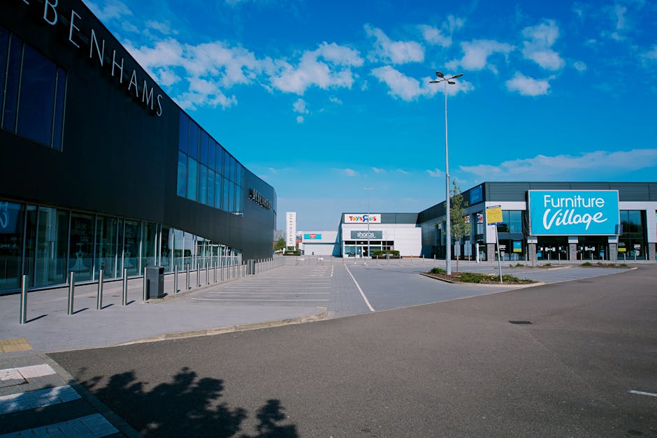 An empty parking lot situated in front of a modern retail complex with multiple stores, including a large black building on the left with the word 'Benhams' visible on its façade, and a blue sign on a grey building on the right advertising 'Furniture Village.' The paved area is largely vacant, with clearly marked parking spaces and a few small patches of greenery with young trees and grass. A tall street lamp stands in the middle of the scene, casting shadows over the asphalt. The sky overhead is bright blue with scattered white clouds, suggesting a clear day. The shopping centre in Lewisham is depicted from a wide-angle perspective, offering a neutral, factual view of the commercial environment, which occasionally relates to non-local authority waste management by highlighting the setting where commercial waste collection might occur, relevant to private waste handling services like those offered by Waste Clearance Lewisham.