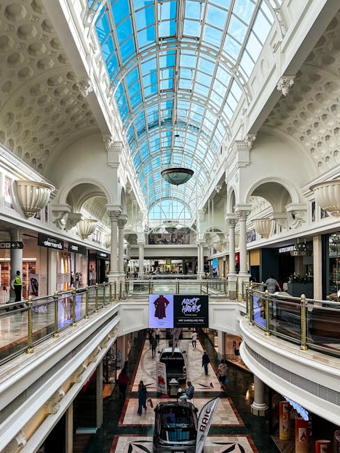 The interior of a modern shopping centre featuring a spacious, multi-level layout with ornate classical architectural elements. The upper balcony is lined with glass and brass railings, overlooking the lower level, which has a polished marble floor with a geometric pattern. Along the sides, storefronts with illuminated signage are visible, with some customers browsing or walking through the mall. The focal point is a large, curved glass ceiling arching overhead, allowing natural daylight to fill the space, supported by decorative white columns with intricate detailing. A digital display screen on the lower level shows an image of a purple garment with promotional text. The environment appears bright and clean, with a calm shopping atmosphere. This setting illustrates an indoor retail environment that prioritizes aesthetic appeal and natural lighting, somewhat akin to private or alternative waste handling spaces with open, well-illuminated interiors. The description is based solely on what is visible and adheres to a neutral and professional tone relevant to the context of waste management and rubbish removal services, where such large commercial spaces often require regular cleaning and waste disposal solutions.