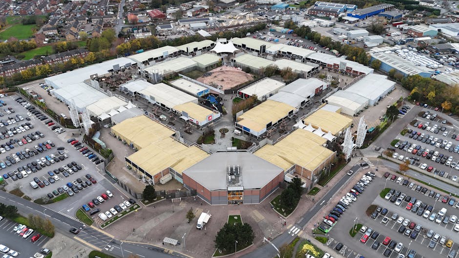 An empty parking lot situated in front of a modern retail complex with multiple stores, including a large black building on the left with the word 'Benhams' visible on its façade, and a blue sign on a grey building on the right advertising 'Furniture Village.' The paved area is largely vacant, with clearly marked parking spaces and a few small patches of greenery with young trees and grass. A tall street lamp stands in the middle of the scene, casting shadows over the asphalt. The sky overhead is bright blue with scattered white clouds, suggesting a clear day. The shopping centre in Lewisham is depicted from a wide-angle perspective, offering a neutral, factual view of the commercial environment, which occasionally relates to non-local authority waste management by highlighting the setting where commercial waste collection might occur, relevant to private waste handling services like those offered by Waste Clearance Lewisham.