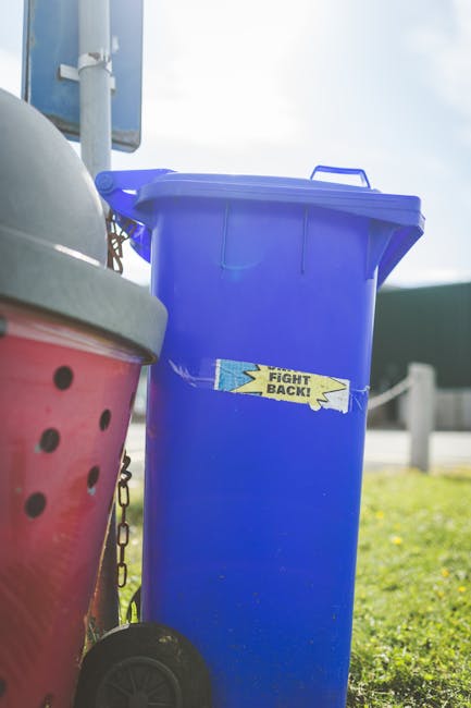 A tall, blue plastic rubbish bin with a closed lid is positioned outdoors on a grassy area, with a small sticker reading 'FIGHT BACK!' affixed to its front. To the left of the bin, a partially visible red container with a black polka dot pattern, possibly a smaller waste or recycling bin, is seen, featuring a gray lid. In the background, a metallic pole and a blue sign are visible, along with a clear sky that indicates daytime. The setting appears to be a public or residential outdoor space, where private waste collection or rubbish disposal services may be involved, aligning with the context of alternative waste handling. The scene is illuminated by natural daylight, providing sharp shadows and vibrant colours, and the environment suggests a typical rubbish collection scene with an emphasis on proper waste segregation and disposal, supported by Waste Clearance Lewisham's professional waste removal services. The overall composition emphasizes the importance of organised rubbish management in community spaces.