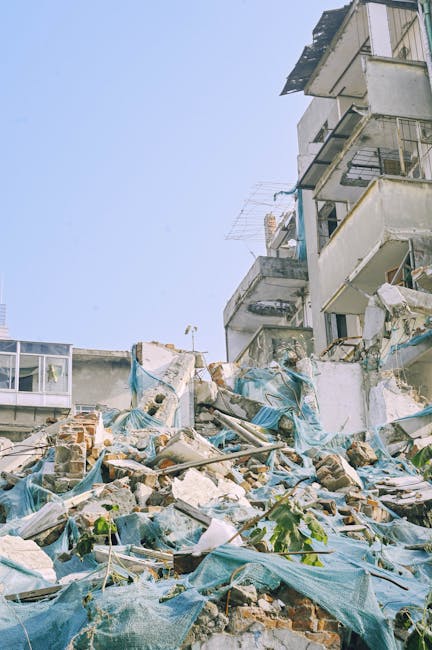 A large pile of demolition debris and construction waste, including broken bricks, concrete fragments, and discarded blue tarpaulins, is situated at the base of a partially collapsed multi-storey building in an urban environment. The debris covers a wide area of the ground and appears to be mixed in irregular heaps, with some bricks and rubble leaning against the remaining building structure on the right side of the image. The building features balconies with metal railings, some of which have visible damage or disrepair, and the exterior walls are a combination of painted and exposed concrete surfaces. The scene is well-lit with natural daylight, and the clear sky overhead provides a neutral backdrop. This scene exemplifies the type of site where professional waste removal services, such as those provided by Waste Clearance Lewisham, are often employed to manage bulky and hazardous construction or demolition waste in an efficient and safe manner, facilitating alternative or independent disposal options beyond standard municipal collection.