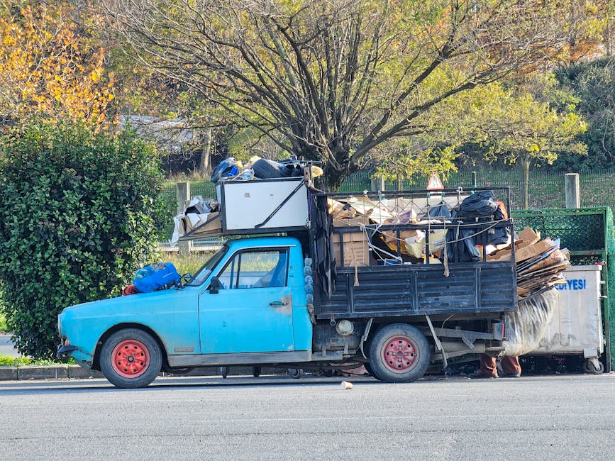 A small, blue, aged pickup truck parked alongside a curb on a street, heavily loaded with assorted waste materials including cardboard boxes, plastic bags, and unusable furniture, which are stacked and secured with ropes on the truck bed and attached to its sides. The truck's body shows visible rust and paint peeling, with red rims on the front wheel, and the driver's side door is closed. In the background, there is a large tree with green and yellow leaves indicating an autumn setting, and a green metal fence runs behind the vehicle. The surrounding environment appears to be a residential or park area with natural greenery and a clear, bright sky. The scene reflects a private waste collection or on-site clearance activity, typical of services offered by waste clearance companies like Waste Clearance Lewisham, especially when managing bulky waste that cannot be disposed of through regular rubbish collection.