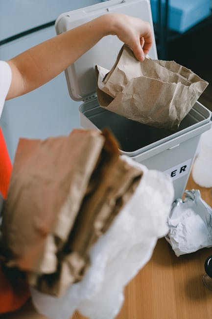 A person’s hand is shown placing a crumpled brown paper bag into a grey composter bin with a partially open lid, situated indoors on a wooden surface. The bin is labeled with partially visible text indicating it is designated for waste collection. In the foreground, several more folded and crumpled paper bags are being held, ready to be disposed of. The background features a light-colored wall and a window, providing natural illumination. The scene highlights a typical household waste disposal process, with emphasis on paper waste suitable for recycling or alternative waste handling services, consistent with the context of rubbish removal and private waste collection options offered by Waste Clearance Lewisham.