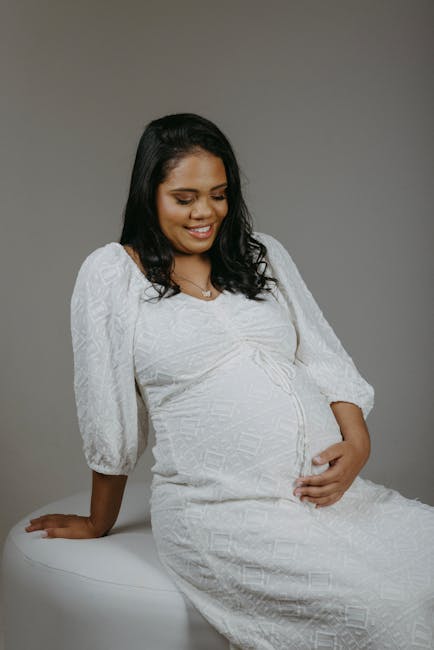 A pregnant woman with medium skin tone and long, dark hair is seated on a white, curved surface against a plain grey background. She is wearing a white textured dress with three-quarter length sleeves and a modest neckline, and she is smiling softly while gently resting her right hand on her baby bump and her left hand on the surface. The smooth fabric of the dress features subtle geometric patterns, and her relaxed posture emphasizes her pregnancy. The neutral lighting highlights her facial features and the soft texture of her clothing. This image appears unrelated to rubbish removal services or waste management and depicts a serene, personal moment in an indoor setting.